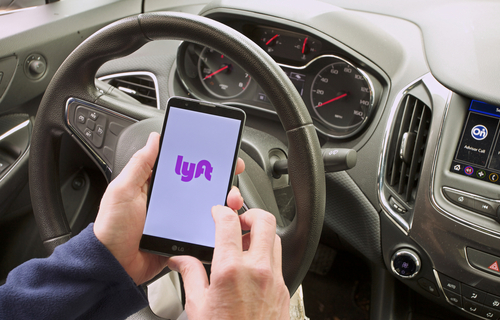 A driver sitting behind the wheel of a car holding a smartphone displaying the Lyft app on the screen, preparing for a rideshare trip.