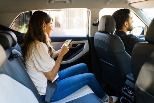 A woman sitting in the back seat of a car, wearing a seatbelt and smiling while using her smartphone, with the driver focused on the road ahead.