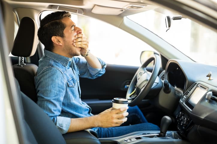 tired man sitting in car yawning and holding a cup of coffee