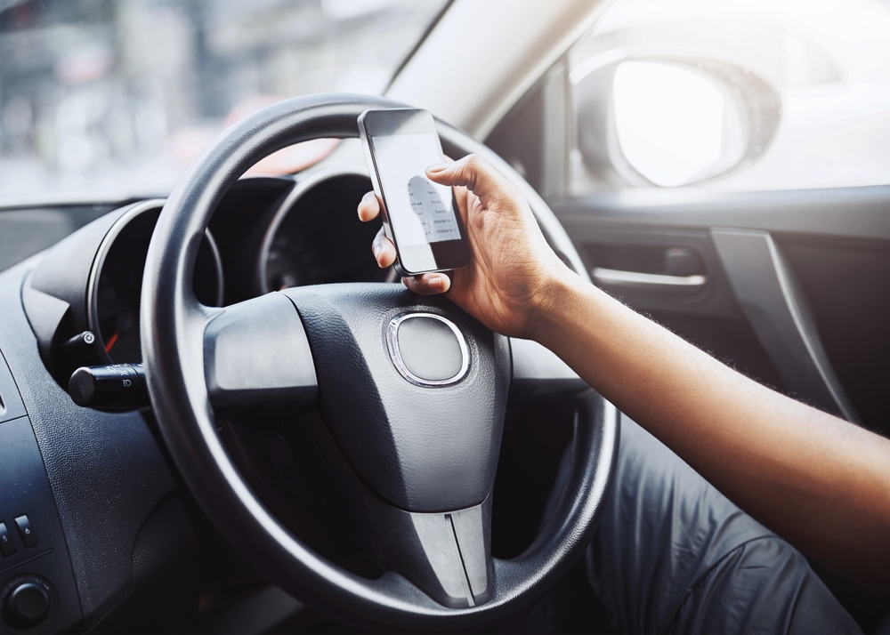 Person texting while driving, holding phone on the steering wheel. Danger and risk. Road safety awareness with driver using smartphone in car.