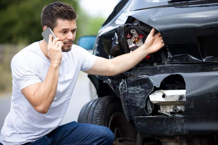 Guy on phone crouching down while holding on to damaged car