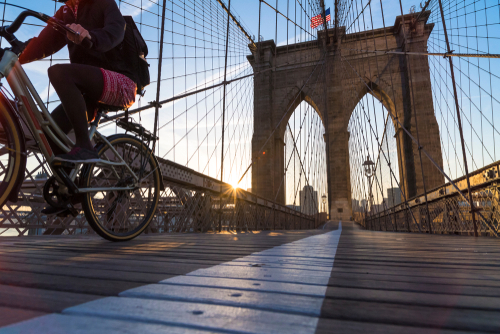 Cyclist riding across the Brooklyn Bridge bike lane in New York City during sunrise.