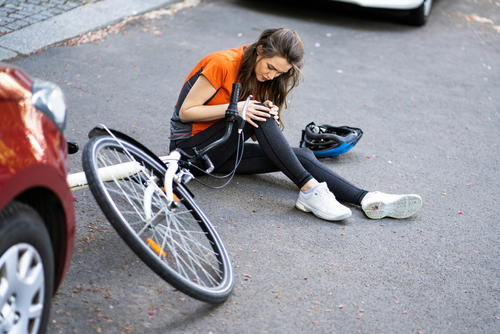 Woman sitting on the road holding her injured knee beside a fallen bicycle after a collision with a red car.