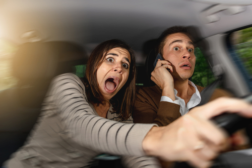 A shocked passenger reaches for the steering wheel while the driver uses a phone, illustrating distracted driving danger.