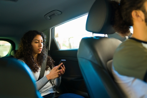 A rideshare passenger looks at her phone in the back seat while the driver focuses on the road ahead.
