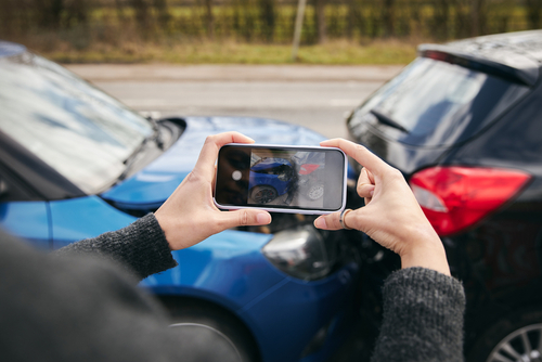 Close-up of a person holding a smartphone and taking photos of damage after a rear-end car accident between a blue and a black vehicle.