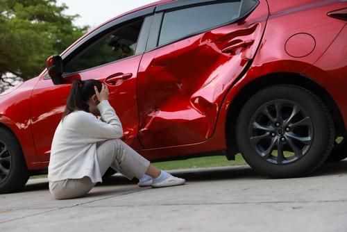 Young woman sitting on the ground next to a red car with a large side dent after a collision, holding her head in distress.