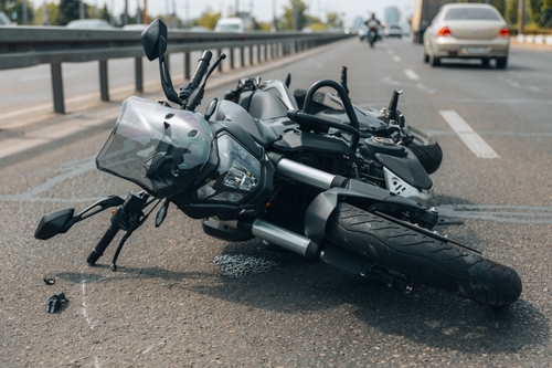 A damaged black motorcycle lying on its side in the middle of a multi-lane highway after an accident, with other vehicles visible in the background.