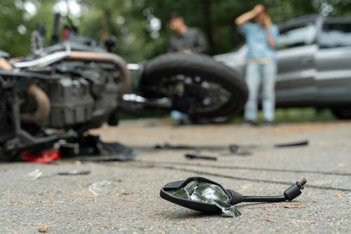 A broken motorcycle mirror and debris scattered on the road after a crash, with a motorcycle and a car involved in the background and two people standing nearby.